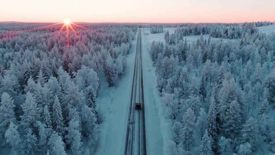 An aerial view of Arctic Lumo Resort's glass-roofed cabins beside the snow-covered forest at sunrise.